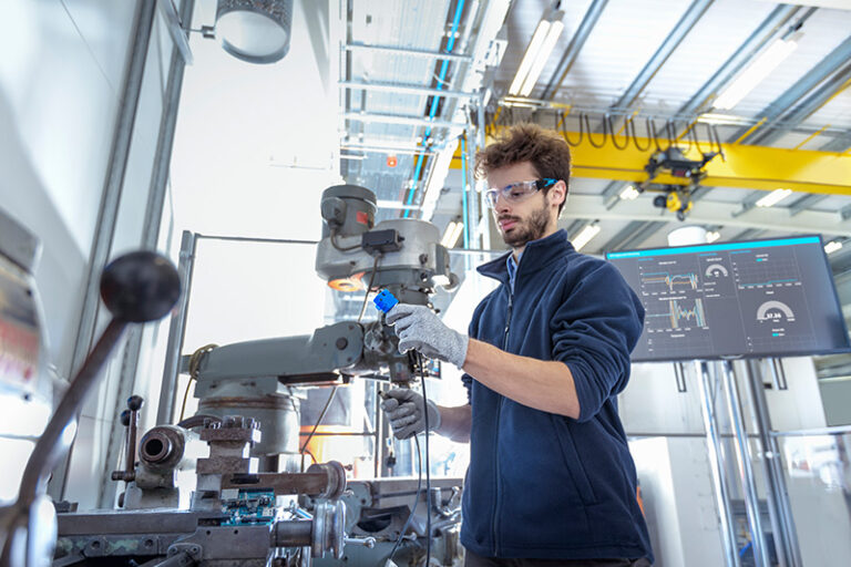 A worker wearing safety glasses and gloves operates machinery in a bright industrial setting. A digital screen with data is visible in the background.