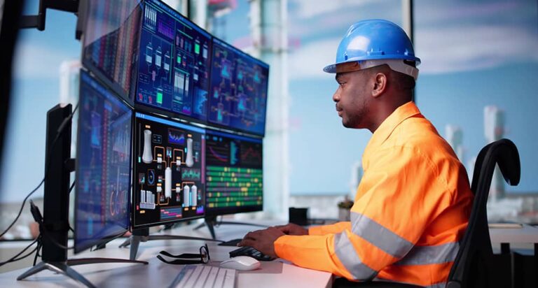 A man in an orange safety jacket and blue hard hat monitors complex data on multiple screens in a control room, conveying focus and precision.