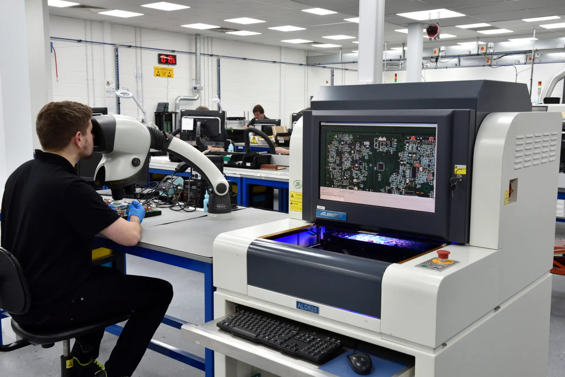 A technician in a black shirt works on electronic equipment at a desk, using a large microscope. Nearby, a screen displays a circuit board layout, indicating a busy and high-tech laboratory environment focused on precision and detail.