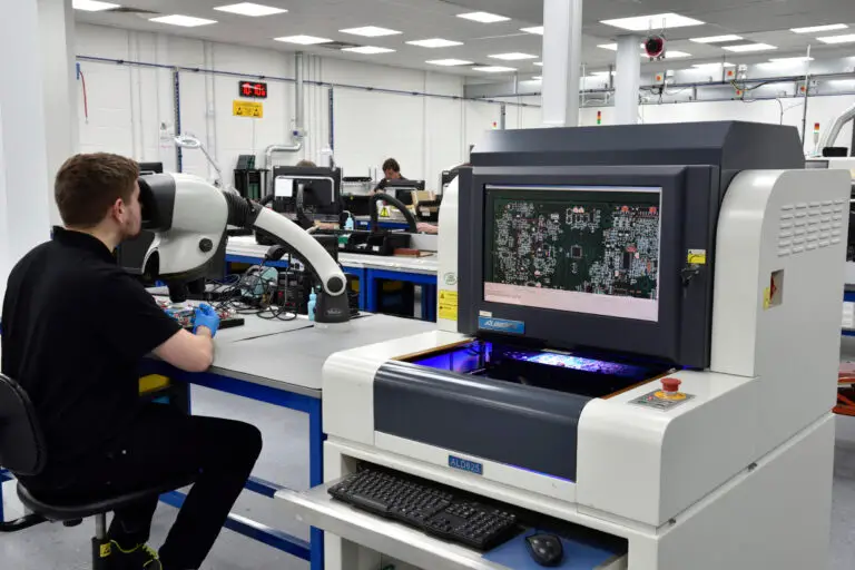 A technician in a black shirt works on electronic equipment at a desk, using a large microscope. Nearby, a screen displays a circuit board layout, indicating a busy and high-tech laboratory environment focused on precision and detail.