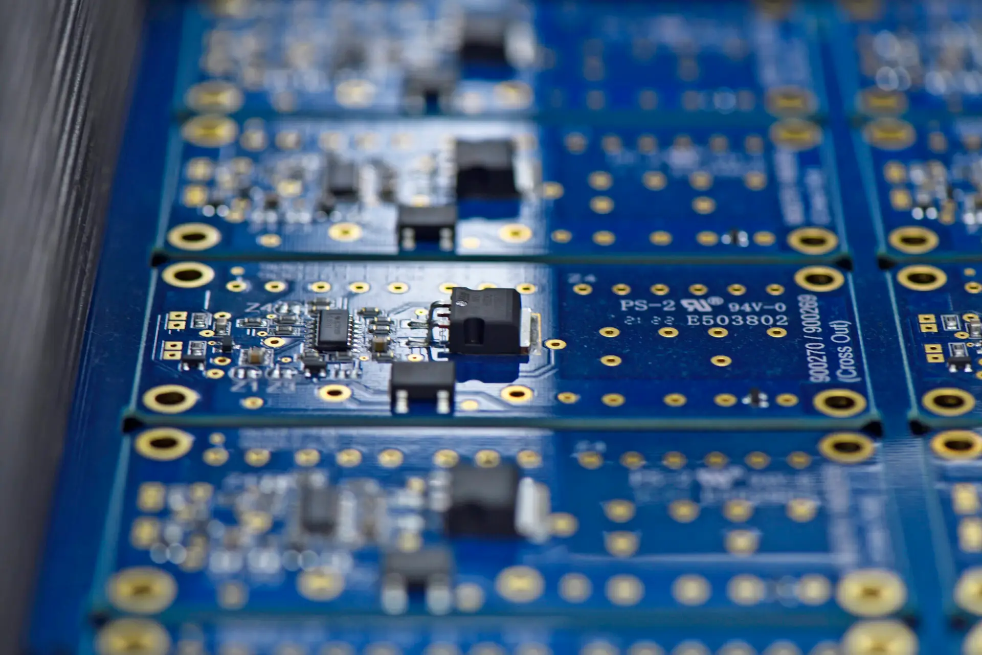 Close-up of several blue printed circuit boards aligned in rows, featuring small electronic components and gold-coloured connections. The tone is technical and precise.