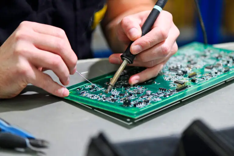 Close-up of hands soldering components on a green circuit board. The scene shows precision and concentration, with tools visible on a workbench.
