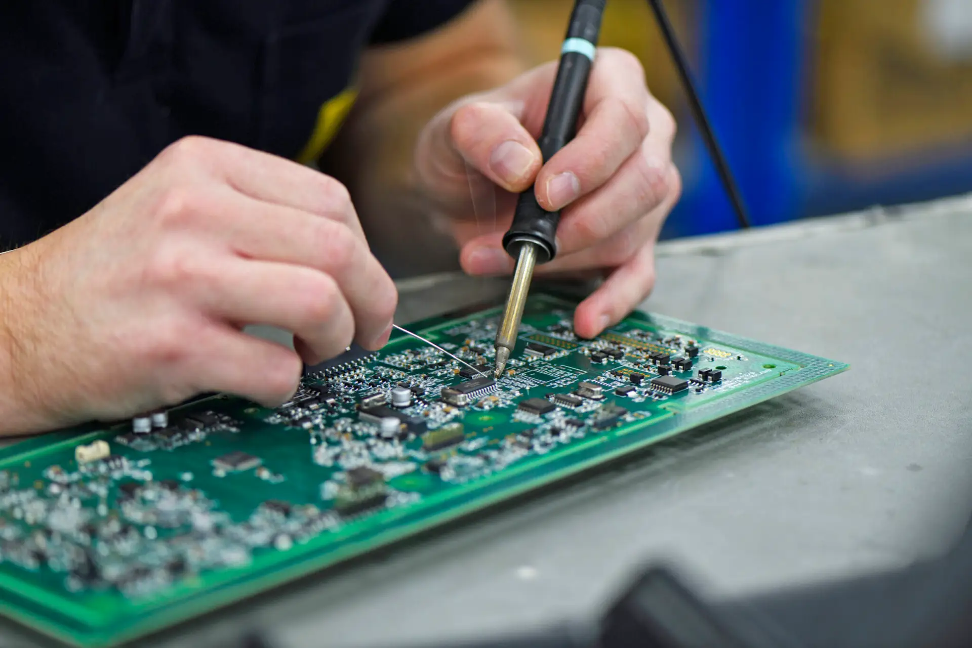 Close-up of hands soldering components on a green circuit board. The scene shows precision and concentration, with tools visible on a workbench.