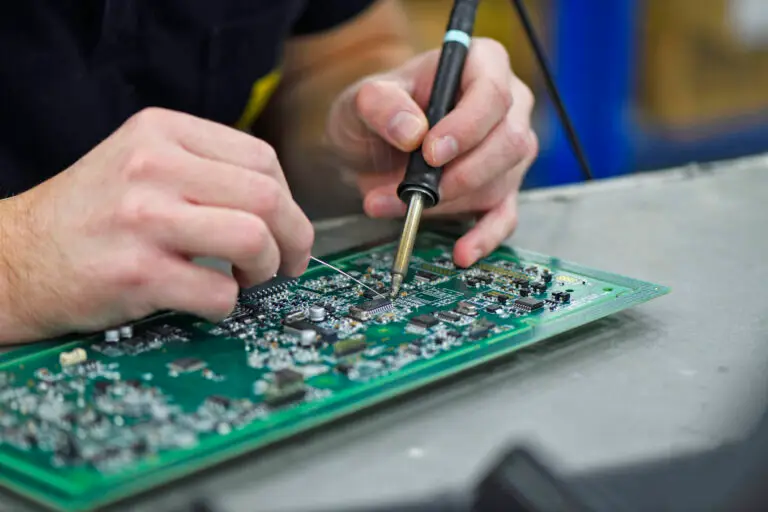 Close-up of hands soldering components on a green circuit board. The scene shows precision and concentration, with tools visible on a workbench.