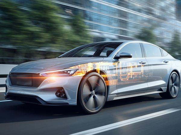 Sleek silver car driving on a city road with holographic technology overlays, suggesting advanced digital features. Background shows motion-blurred trees.