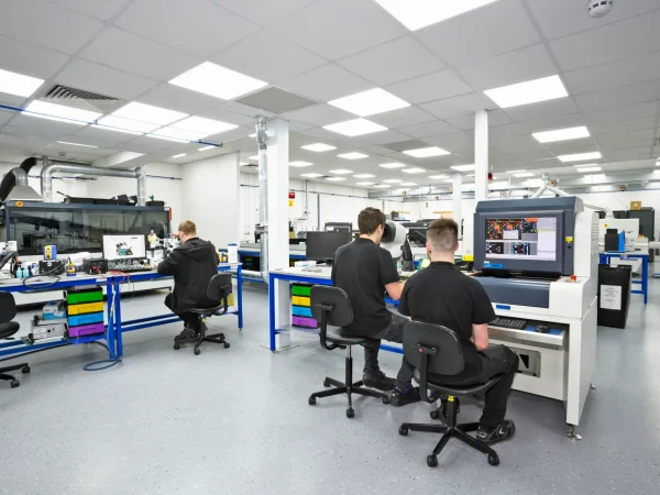 A modern electronics lab with bright lighting, three technicians at workstations focused on circuit diagrams and screens. The scene conveys focus and innovation.