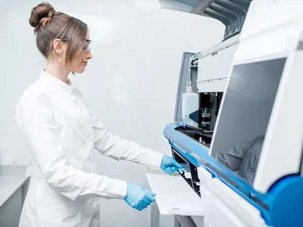 A scientist in a white lab coat and safety glasses uses a large laboratory machine. She wears blue gloves, conveying a professional, focused atmosphere.
