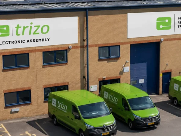 Trizo Electronic Assembly facility with rooftop solar panels, brick walls, and three bright green branded vans parked outside on a sunny day.