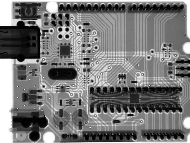 X-ray image of a circuit board showing intricate wiring and components, including a USB port on the left. The black, white, and gray tones reveal fine details.