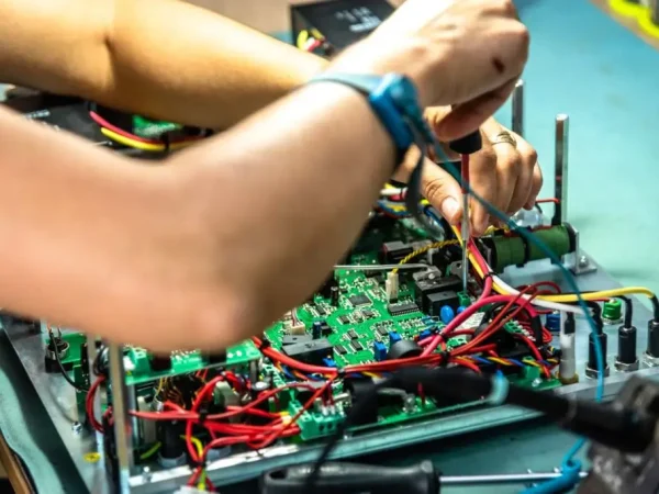 Person working on a circuit board with various tools, surrounded by colourful wires. The setting conveys a focused, technical atmosphere.